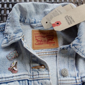Puede incluir: Chaqueta vaquera azul claro con una etiqueta de Levi's y una pequeña etiqueta que dice "Original Trucker Jacket". La chaqueta tiene cierre de botones y dos bolsillos en la parte delantera. Hay dos pines en la chaqueta, uno que dice "Dope" y otro que es un patín.