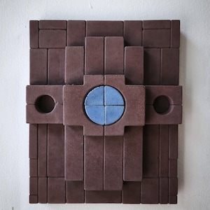 Könnte beinhalten: Geometrische Wandkunst aus rechteckigen und kreisförmigen Formen in Braun- und Blautönen. Das Design zeigt einen zentralen blauen Kreis und zwei kreisförmige Aussparungen, die eine moderne, abstrakte Ästhetik schaffen. Das Kunstwerk ist an einer weißen Wand befestigt.