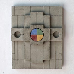 Puede incluir: Una obra de arte mural de hormigón gris con un diseño geométrico. La obra presenta un elemento circular central dividido en cuatro secciones de color: azul, verde, rojo y amarillo. Dos agujeros circulares están a cada lado del elemento central.