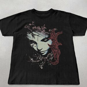 K&ouml;nnte beinhalten: Schwarzes T-Shirt mit einem stilisierten Gesicht, floralen Elementen und einem roten, verzierten Design. Das Gesicht hat helle Haut und dunkles Haar. Das Design befindet sich auf der Vorderseite des Shirts.