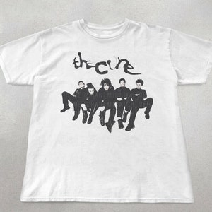 May include: White t-shirt with a black and white image of the band The Cure. The band members are sitting and the band's name is written in a stylized font above them.
