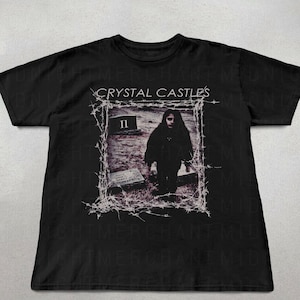 Könnte beinhalten: Schwarzes T-Shirt mit dem Bandnamen "Crystal Castles" über einem gerahmten Bild einer Person auf einem Friedhof. Das Bild enthält einen Grabstein und die Zahl "II". Das Design ist von einem weißen, stacheldrahtartigen Rand umgeben.