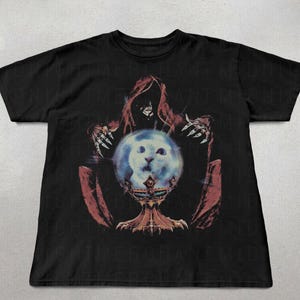 May include: Black t-shirt featuring a graphic of a hooded figure with clawed hands holding a crystal ball with a cat's face inside. The crystal ball sits on a decorative stand. The design is in shades of red, blue, and gold.