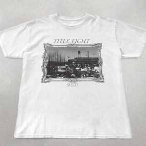 Könnte beinhalten: Weißes T-Shirt mit einem Schwarzweißbild eines Gebäudes in einem Rahmen. Der Text "TITLE FIGHT" und "SHED" ist auf dem Shirt aufgedruckt.