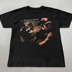 Puede incluir: Camiseta negra con la inscripción "Elliott Smith either/or" y una imagen descolorida de una persona con gorra de béisbol. El diseño tiene un aspecto vintage y ligeramente desgastado.