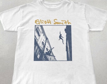 Puede incluir: Camiseta blanca con una imagen azul y blanca de una persona cayendo de un edificio. El texto "Elliott Smith" está escrito en dorado sobre la imagen.
