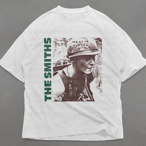 Puede incluir: Camiseta blanca con un estampado verde y marr&oacute;n de una persona con un casco con el texto "Meat is Murder". El texto "The Smiths" est&aacute; impreso verticalmente en el lado izquierdo de la camiseta.