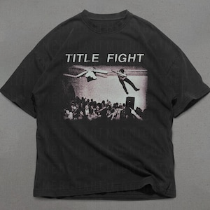 Op de afbeelding: Donkergrijs t-shirt met een grafische print. De print toont de woorden "TITLE FIGHT" boven een zwart-wit afbeelding van een persoon die van een podium in een menigte valt. Het shirt heeft korte mouwen en een ronde hals.
