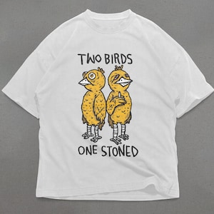 Pode incluir: Camiseta branca com um gráfico de dois pássaros amarelos, um com um cigarro no bico. O texto "TWO BIRDS ONE STONED" está impresso abaixo dos pássaros.