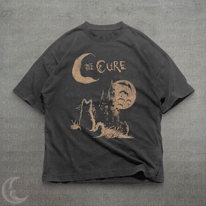 May include: A charcoal gray short-sleeve t-shirt featuring a tan graphic print of a cat sitting before a castle, a crescent moon, and a full moon. The text "THE CURE" is printed in a stylized font above the cat and castle illustration.