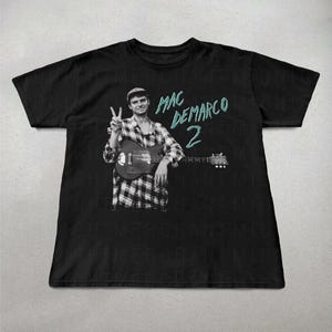 May include: Black t-shirt featuring a black and white image of a person holding a guitar and giving a peace sign. The text "MAC DEMARCO 2" is in teal. The person is wearing a plaid shirt and a cap. The shirt is a crew neck.