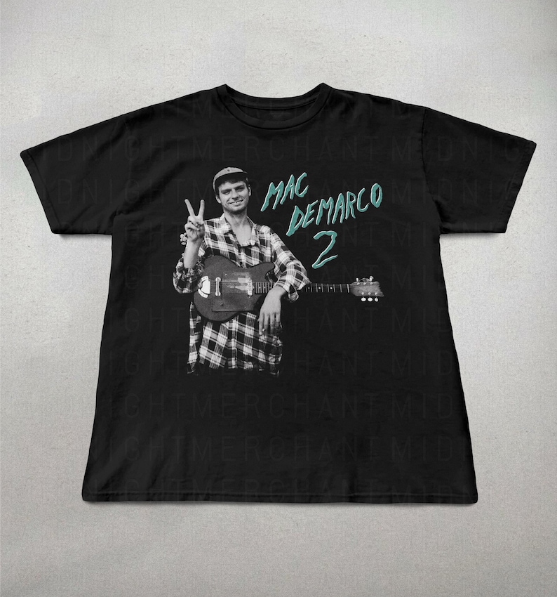 May include: Black t-shirt featuring a black and white image of a person holding a guitar and giving a peace sign. The text "MAC DEMARCO 2" is in teal. The person is wearing a plaid shirt and a cap. The shirt is a crew neck.