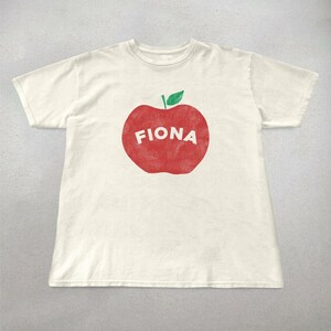 May include: Off-white t-shirt featuring a large red apple graphic with the name "FIONA" in white block letters. The apple has a green leaf at the top. The shirt is a short-sleeved crew neck.