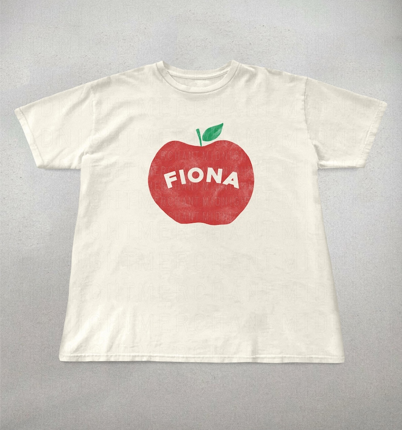 May include: Off-white t-shirt featuring a large red apple graphic with the name "FIONA" in white block letters. The apple has a green leaf at the top. The shirt is a short-sleeved crew neck.