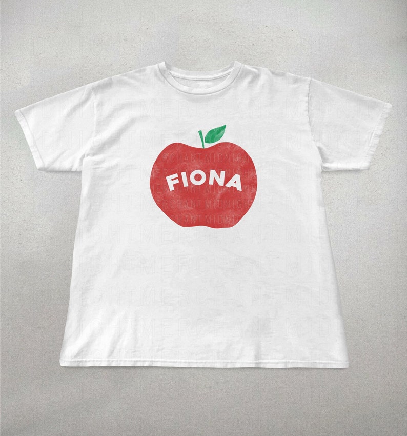 May include: White t-shirt featuring a large red apple graphic with the name "FIONA" in white letters. The apple has a green leaf at the top. The shirt is made of a soft, comfortable material.