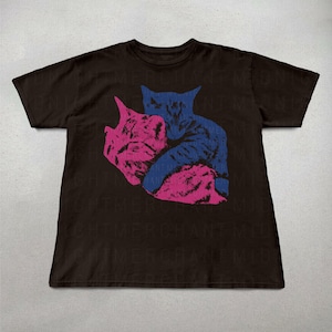 Pu&ograve; includere: T-shirt marrone scuro con grafica di due gatti che si abbracciano. Un gatto &egrave; rosa e l'altro blu. Il disegno &egrave; stampato sul davanti. La maglietta ha maniche corte e girocollo.
