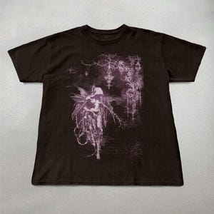 May include: Dark brown t-shirt featuring a detailed, gothic-style illustration in shades of purple and white. The design includes a figure with wings and intricate, ornamental elements. The shirt is made of a soft material.