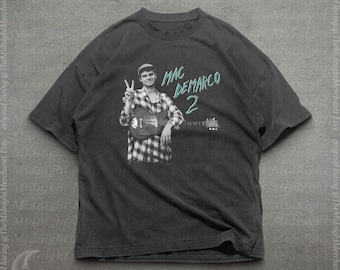 Op de afbeelding: Een antracietgrijs T-shirt met korte mouwen en een grafische print van een persoon in een geruit overhemd die een elektrische gitaar vasthoudt en een vredesteken maakt. De tekst "MAC DEMARCO 2" staat in lichtblauwe letters naast de figuur.