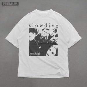 Puede incluir: Camiseta blanca con una imagen en blanco y negro de una banda, la palabra "slowdive" arriba y "souvlaki" abajo. La camiseta tiene cuello redondo y mangas cortas.