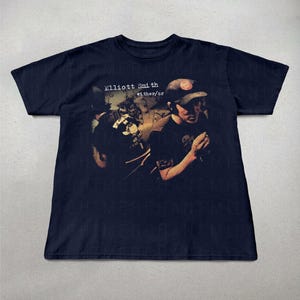 May include: Navy blue t-shirt with the text "Elliott Smith either/or" and a faded image of a person wearing a baseball cap. The graphic design is in shades of brown and black.