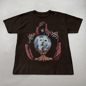 May include: Dark brown t-shirt featuring a graphic design. The design depicts a hooded figure with outstretched hands, holding a glowing orb with a cat's face. The orb rests on a decorative chalice. The design is in shades of red, blue, and white.