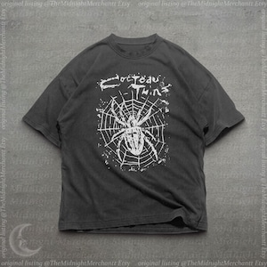 Pode incluir: Uma t-shirt cinza-carvão de manga curta com uma estampa gráfica branca de uma aranha numa teia. Acima da aranha, o texto diz Cocteau Twins numa fonte estilizada e desgastada. A t-shirt tem uma gola redonda.