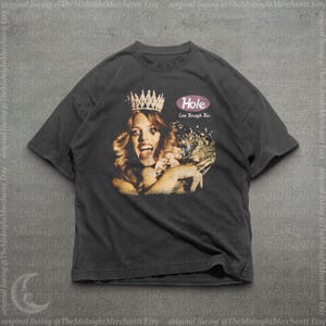 May include: A charcoal gray short-sleeve t-shirt featuring a graphic print of a person wearing a crown and holding flowers. The text "Hole" appears in a pink oval above the words "Live Through This" in a stylized white font on the chest.