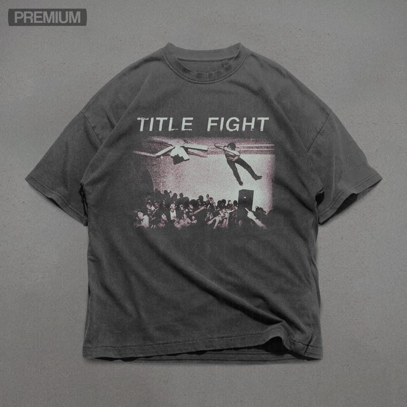 Op de afbeelding: Donkergrijs T-shirt met de tekst "TITLE FIGHT" boven een afbeelding van een persoon die van een podium in een menigte valt. Het shirt heeft korte mouwen en een ronde hals.