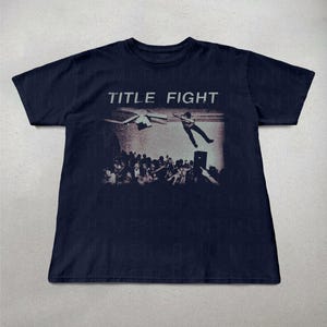 May include: Navy blue t-shirt featuring a black and white graphic of a concert scene with the text "TITLE FIGHT" across the top. The graphic shows a person jumping off a stage and a crowd of people.