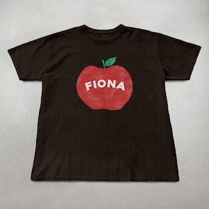 May include: A dark brown t-shirt featuring a large red apple graphic with the name "FIONA" in white letters. The apple has a green leaf at the top. The shirt is a casual style.