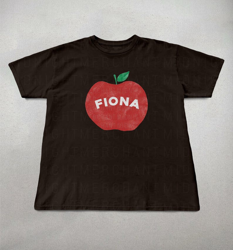 May include: A dark brown t-shirt featuring a large red apple graphic with the name "FIONA" in white letters. The apple has a green leaf at the top. The shirt is a casual style.