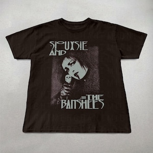 May include: Dark brown t-shirt featuring a black and white image of Siouxsie Sioux holding a skull, with the band name "Siouxsie and the Banshees" printed above and below the image in a gothic font.