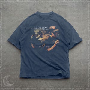 May include: A dark blue short-sleeve t-shirt featuring a graphic print of the Elliott Smith either/or album cover. The design shows a person wearing a baseball cap, with the text Elliott Smith either/or printed in white above the image.