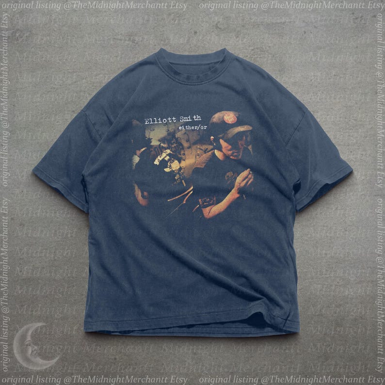 May include: A dark blue short-sleeve t-shirt featuring a graphic print of the Elliott Smith either/or album cover. The design shows a person wearing a baseball cap, with the text Elliott Smith either/or printed in white above the image.