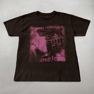 May include: A dark brown t-shirt featuring a pink graphic with the text "my bloody valentine" and "loveless". The graphic appears to be a blurred image of a guitar. The shirt is a short-sleeved crew neck.
