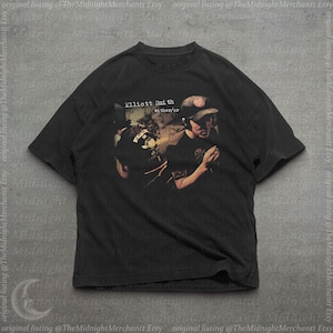 May include: A black short-sleeve t-shirt featuring a graphic print of a person wearing a cap and singing into a microphone. The text "Elliott Smith" and "either/or" is printed above the figure in a white, slightly distressed, serif font.