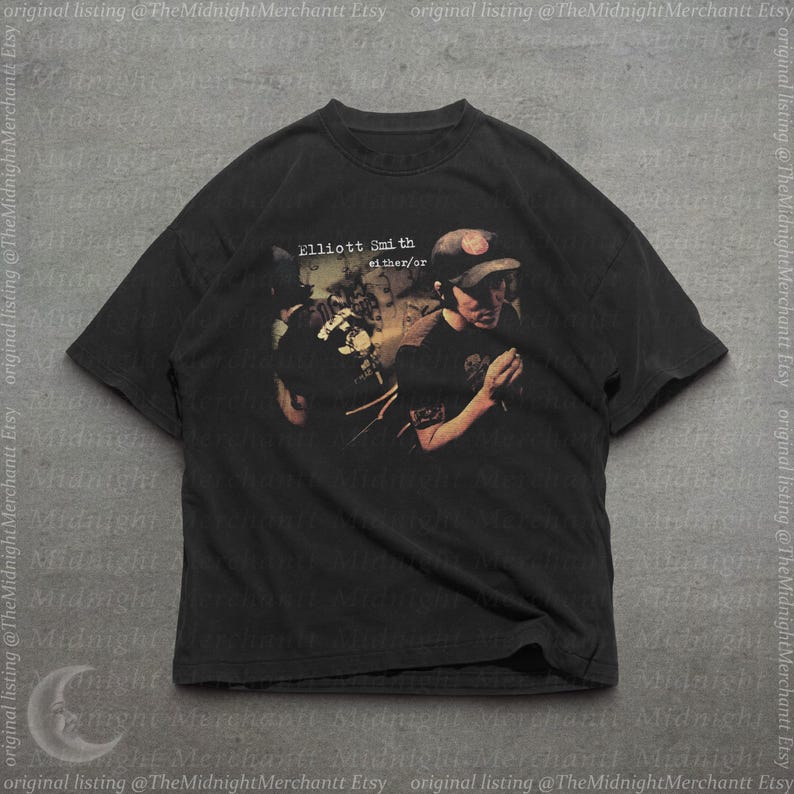 May include: A black short-sleeve t-shirt featuring a graphic print of a person wearing a cap and singing into a microphone. The text "Elliott Smith" and "either/or" is printed above the figure in a white, slightly distressed, serif font.
