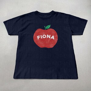 May include: Navy blue t-shirt featuring a large red apple graphic with the name "FIONA" in white block letters. The apple has a green leaf. The shirt is a casual wear item.