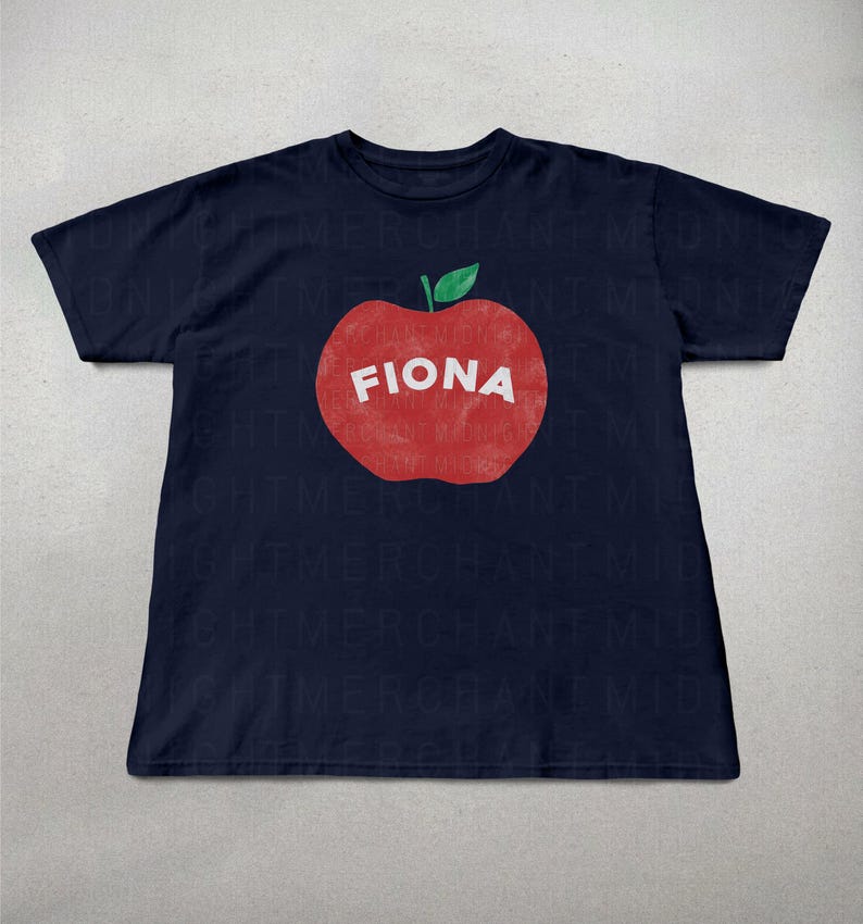 May include: Navy blue t-shirt featuring a large red apple graphic with the name "FIONA" in white block letters. The apple has a green leaf. The shirt is a casual wear item.