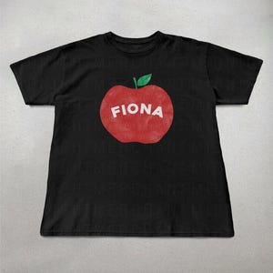 May include: Black t-shirt featuring a large red apple graphic with the name "FIONA" in white block letters. The apple has a green leaf at the top. The shirt is made of a soft material.
