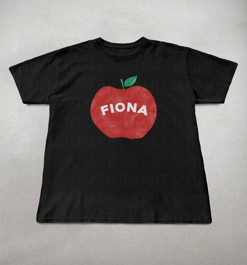 May include: Black t-shirt featuring a large red apple graphic with the name "FIONA" in white block letters. The apple has a green leaf at the top. The shirt is made of a soft material.