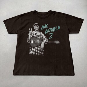 May include: Black t-shirt featuring a black and white image of a person holding a guitar and giving a peace sign. The text "MAC DEMARCO 2" is printed in teal on the shirt.