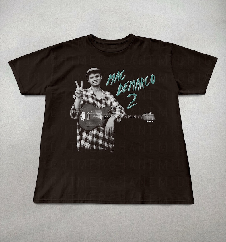 May include: Black t-shirt featuring a black and white image of a person holding a guitar and giving a peace sign. The text "MAC DEMARCO 2" is printed in teal on the shirt.