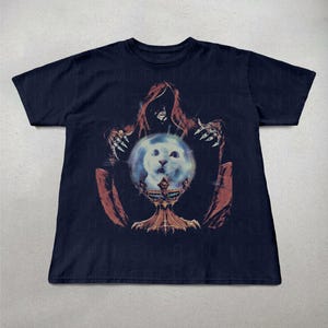 May include: Navy blue t-shirt featuring a graphic of a hooded figure with clawed hands, holding a glowing orb with a cat's face. The orb rests on a decorative chalice. The design is in shades of red, orange, and blue.