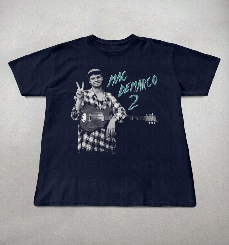 May include: Navy blue t-shirt featuring a black and white image of a musician holding a guitar and making a peace sign. The text "MAC DEMARCO 2" is printed in teal on the shirt. The shirt is a short-sleeved crew neck.