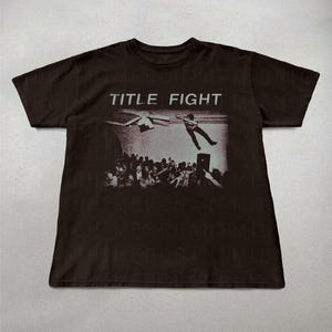 May include: Dark brown t-shirt featuring a black and white graphic of a person jumping over a crowd, with the text "TITLE FIGHT" above the image. The shirt is a crew neck style.