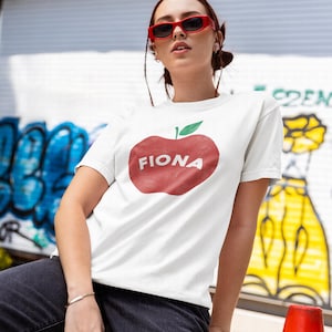 May include: White t-shirt featuring a large red apple graphic with the name "FIONA" in white letters. The apple has a green leaf. The person is wearing red sunglasses. The background has graffiti art.