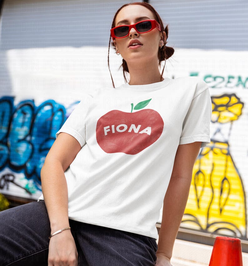 May include: White t-shirt featuring a large red apple graphic with the name "FIONA" in white letters. The apple has a green leaf. The person is wearing red sunglasses. The background has graffiti art.