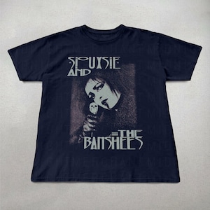 May include: Navy blue t-shirt featuring a graphic of Siouxsie Sioux and the band name, Siouxsie and the Banshees, in white text. The image shows a portrait of Siouxsie holding a skull. The shirt is a classic crew neck style.