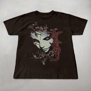 K&ouml;nnte beinhalten: Dunkelbraunes T-Shirt mit einer Grafik eines stilisierten Gesichts, floralen und abstrakten Elementen. Das Gesicht ist in Blau und Wei&szlig; gehalten, mit roten Akzenten. Das Design ist mittig auf der Vorderseite des T-Shirts platziert.